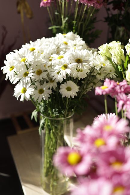 A clear glass vase containing a fresh bouquet of white daisies with yellow centers, arranged with long green stems visible through the transparent glass. The daisies are placed on a light-colored surf