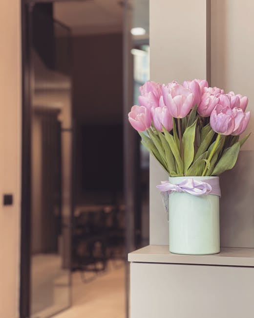 A bouquet of pink tulips with smooth, rounded petals and fresh green leaves is arranged in a light green ceramic vase decorated with a delicate lavender ribbon tied in a bow. The flowers are vibrant a