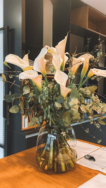 A large, elegant floral arrangement in a clear glass vase features cream-colored calla lilies with smooth, curved petals and long green stems. Interspersed among the calla lilies are delicate sprigs o