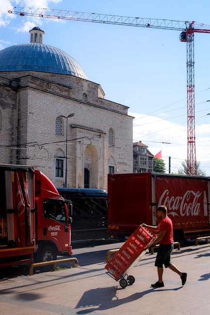 The image features a worker in a red shirt and black shorts unloading multiple red Coca-Cola branded crates onto the sidewalk alongside a Coca-Cola delivery truck. In the background, there is a histor