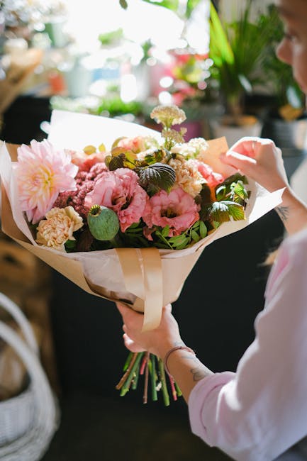 A person holding a vibrant bouquet of fresh flowers, including pink peonies, white hydrangeas, and green foliage, wrapped in elegant white and beige paper with a cream-colored ribbon. The bouquet is p